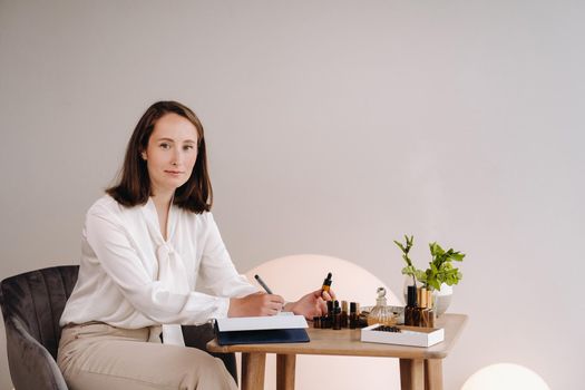 The aromatherapist girl is sitting in her office and holding a bottle of aromatic oil in her hands and writing something down. there are essential oils on the table.