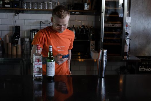 stylish young man hipster in orange t-shirt making mixing a cocktail in a dark loft cafe. alcohol drink in modern bar.