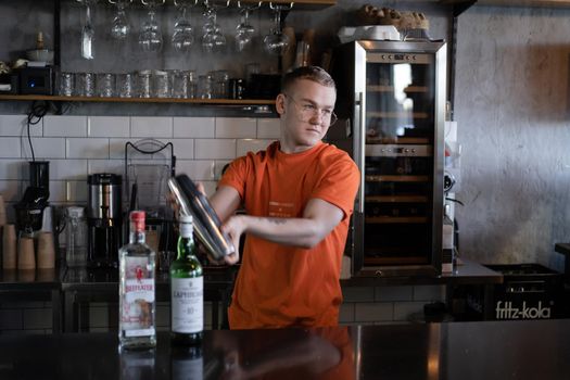 stylish young man hipster in orange t-shirt making mixing a cocktail in a dark loft cafe. alcohol drink in modern bar.