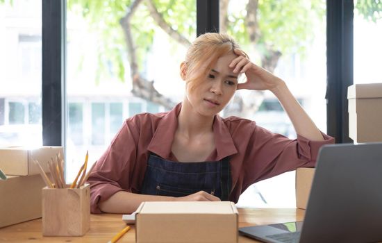 A portrait of young serious Asian woman working with laptop in the office full of packages and boxes stacking up, busy looking table, for SME, delivery, start up business and home office concept..