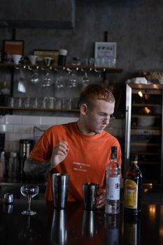 stylish young man hipster in orange t-shirt making mixing a cocktail in a dark loft cafe. alcohol drink in modern bar.