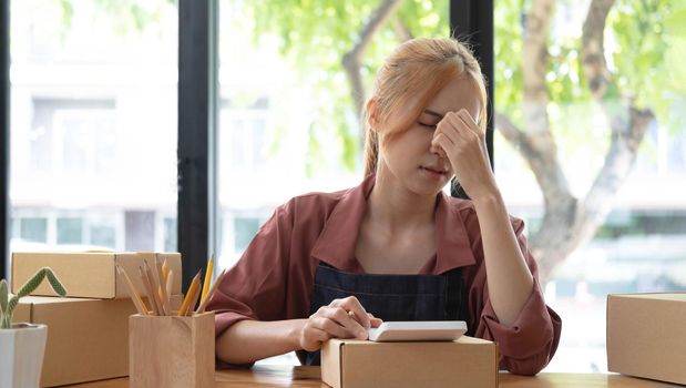 A portrait of young serious Asian woman working with laptop in the office full of packages and boxes stacking up, busy looking table, for SME, delivery, start up business and home office concept.