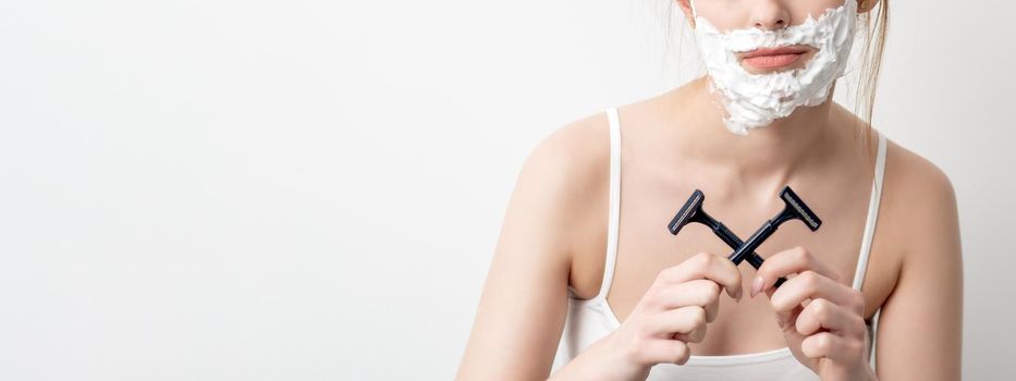 Beautiful young caucasian woman with shaving foam on her face and two razors in her hands on white background