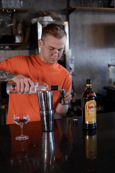 stylish young man hipster in orange t-shirt making mixing a cocktail in a dark loft cafe. alcohol drink in modern bar.