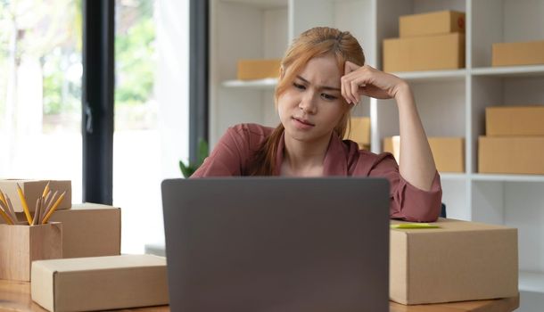 A portrait of young serious Asian woman working with laptop in the office full of packages and boxes stacking up, busy looking table, for SME, delivery, start up business and home office concept.