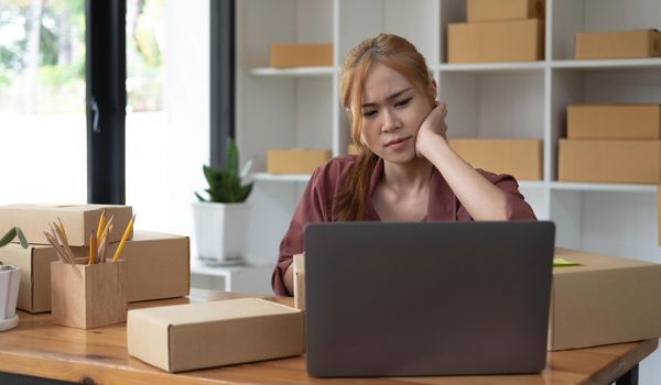 A portrait of young serious Asian woman working with laptop in the office full of packages and boxes stacking up, busy looking table, for SME, delivery, start up business and home office concept.