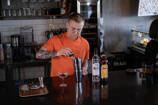 stylish young man hipster in orange t-shirt making mixing a cocktail in a dark loft cafe. alcohol drink in modern bar.