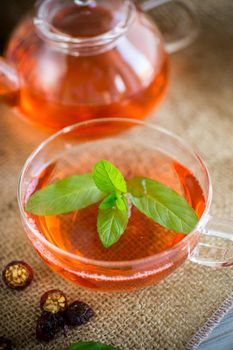 brewed rosehip tea in a glass teapot with rosehip flowers and mint, on a wooden table.