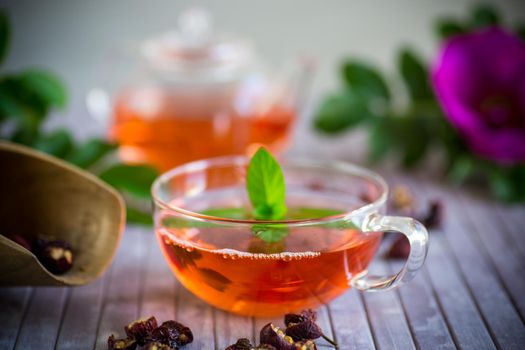 brewed rosehip tea in a glass teapot with rosehip flowers and mint, on a wooden table.