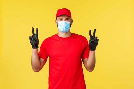 Express delivery during pandemic, covid-19, safe shipping, online shopping concept. Friendly courier in red uniform, medical mask and gloves, show peace sign, yellow background.