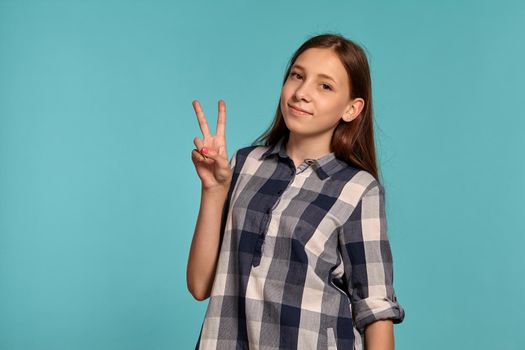 Portrait of a gorgeous teenage female in a casual checkered shirt gesticulating and smiling while posing against a blue studio background. Long hair, healthy clean skin and brown eyes. Sincere emotions concept. Copy space.