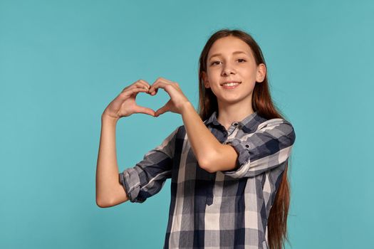 Portrait of a gorgeous teenage girl in a casual checkered shirt showing a heart sign of her folded fingers and posing against a blue studio background. Long hair, healthy clean skin and brown eyes. Sincere emotions concept. Copy space.