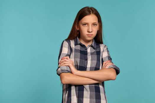Portrait of a charming teenage girl in a casual checkered shirt embraced herself and looking upset while posing against a blue studio background. Long hair, healthy clean skin and brown eyes. Sincere emotions concept. Copy space.