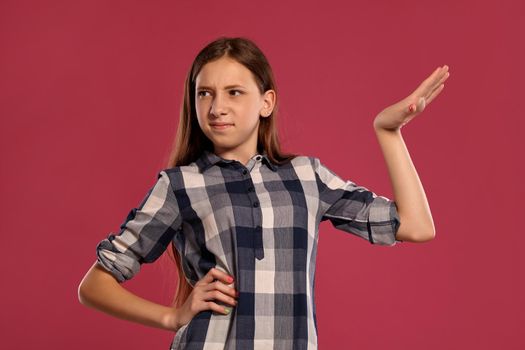Portrait of a beautiful teenage female in a casual checkered shirt looking unsatisfied and posing against a pink studio background. Long hair, healthy clean skin and brown eyes. Sincere emotions concept. Copy space.