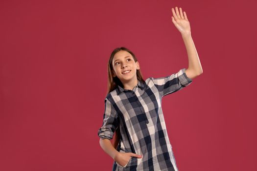 Portrait of an attractive teenage lady in a casual checkered shirt saying hi to someone while posing against a pink studio background. Long hair, healthy clean skin and brown eyes. Sincere emotions concept. Copy space.