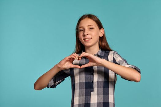 Portrait of a gorgeous teenage lady in a casual checkered shirt showing a heart sign of her folded fingers and smiling while posing against a blue studio background. Long hair, healthy clean skin and brown eyes. Sincere emotions concept. Copy space.