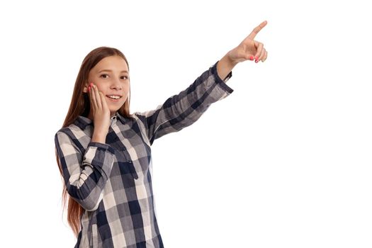 Portrait of a cute teenage girl in a casual checkered shirt pointing at something and looking at the camera while posing isolated on white studio background. Long hair, healthy clean skin and brown eyes. Sincere emotions concept. Copy space.