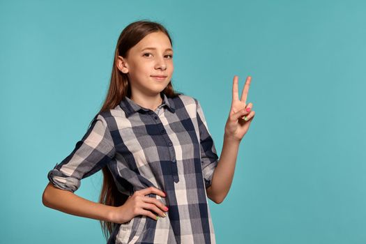 Portrait of a gorgeous teenager in a casual checkered shirt showing a peace sign while posing against a blue studio background. Long hair, healthy clean skin and brown eyes. Sincere emotions concept. Copy space.