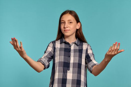 Portrait of a beautiful teenager in a casual checkered shirt gesticulating and posing against a blue studio background. Long hair, healthy clean skin and brown eyes. Sincere emotions concept. Copy space.