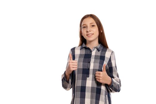 Portrait of an attractive teenage lady in a casual checkered shirt showing thumbs up whie posing isolated on white studio background. Long hair, healthy clean skin and brown eyes. Sincere emotions concept. Copy space.