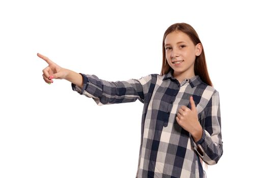 Portrait of a cute teenage maiden in a casual checkered shirt pointing at something and looking at the camera while posing isolated on white studio background. Long hair, healthy clean skin and brown eyes. Sincere emotions concept. Copy space.