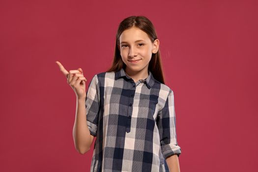 Portrait of a gorgeous teenage girl in a casual checkered shirt gesticulating and looking at the camera while posing against a pink studio background. Long hair, healthy clean skin and brown eyes. Sincere emotions concept. Copy space.