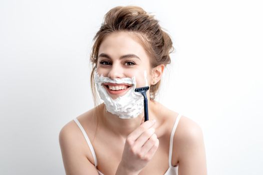 Beautiful young caucasian woman shaving her face by razor on white background. Pretty woman with shaving foam on her face