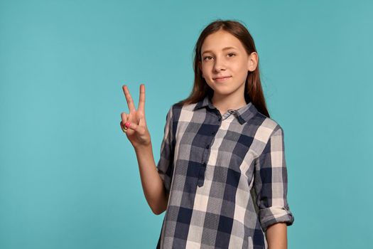 Portrait of an attractive teenage girl in a casual checkered shirt showing two fingers and smiling while posing against a blue studio background. Long hair, healthy clean skin and brown eyes. Sincere emotions concept. Copy space.