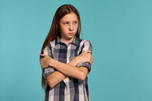 Portrait of a beautiful teenage female in a casual checkered shirt embraced herself and looking offended while posing against a blue studio background. Long hair, healthy clean skin and brown eyes. Sincere emotions concept. Copy space.