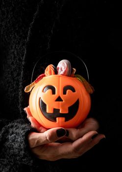 Happy Halloween concept. Scary woman Halloween hand with black nails holding pumpkin full of sweets on dark background with copy space