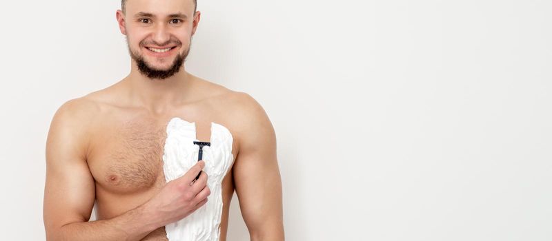 Young caucasian man with beard holds razor shaves his chest with white shaving foam on white background. Man shaving his torso