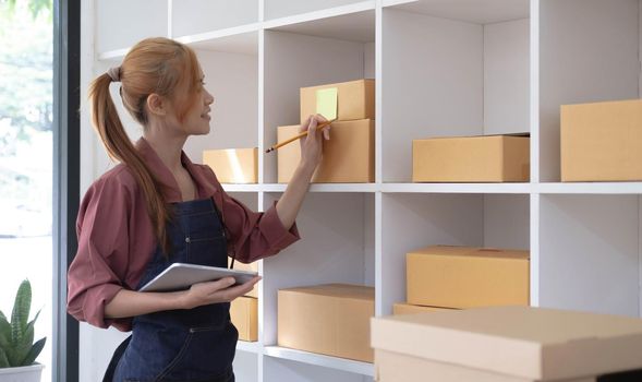 Young business woman working online e-commerce shopping at her shop. Young woman seller prepare parcel box of product for deliver to customer. Online selling, sme, e-commerce..