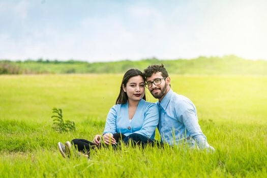 Portrait of a couple in love sitting on the grass in the field, Romantic couple sitting on the grass looking at the camera, Two lovers sitting on the grass looking at the camera