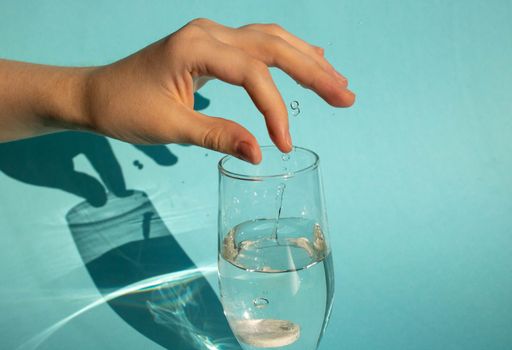 Against a blue background, a hand drops a dissolving fizzy aspirin tablet into a glass of water.