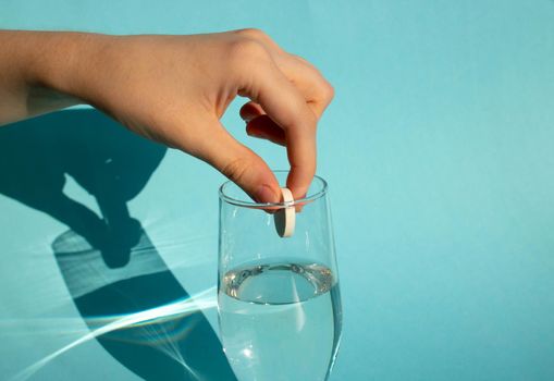 Against a blue background, a hand drops a dissolving fizzy aspirin tablet into a glass of water.