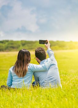 People in love taking selfies in the field with their smartphone, Smiling couple in love sitting on the grass taking selfies, Young couple in love taking a selfie in the field
