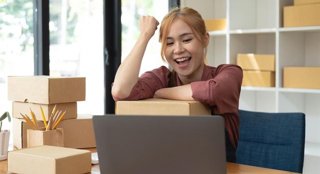 Happy young asian woman startup small business freelance holding parcel box and computer laptop and sitting on chair, Online marketing packing box delivery concept.