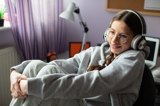 Brunette girl with glasses sits at her room and listens to music. A young girl smiling and full of musical excitement.