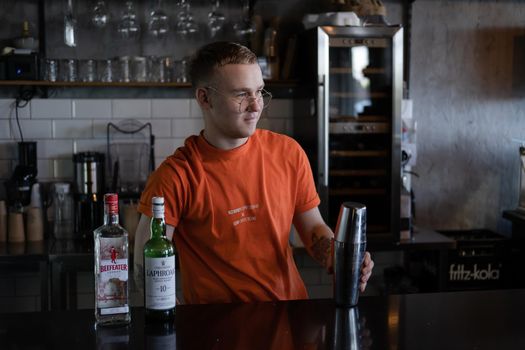 stylish young man hipster in orange t-shirt making mixing a cocktail in a dark loft cafe. alcohol drink in modern bar.