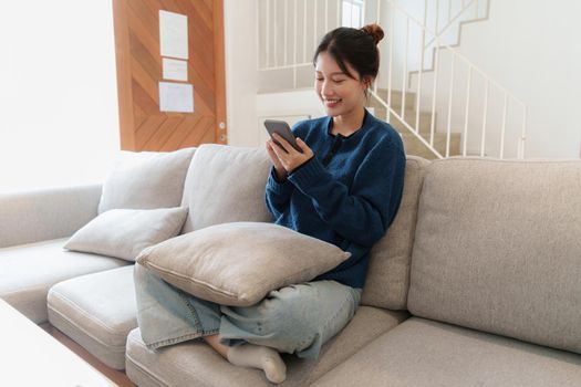 Portrait of Young Asian woman chatting with her friend and checking social media by smartphone sitting on couch. Lifestyle concept