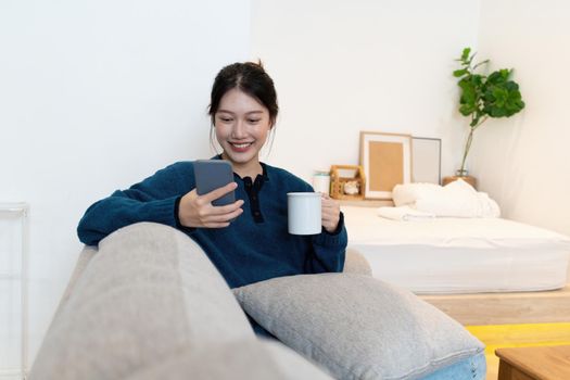 Portrait of Young Asian woman chatting with her friend and checking social media by smartphone sitting on couch. Lifestyle concept