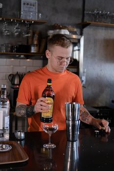 stylish young man hipster in orange t-shirt making mixing a cocktail in a dark loft cafe. alcohol drink in modern bar.