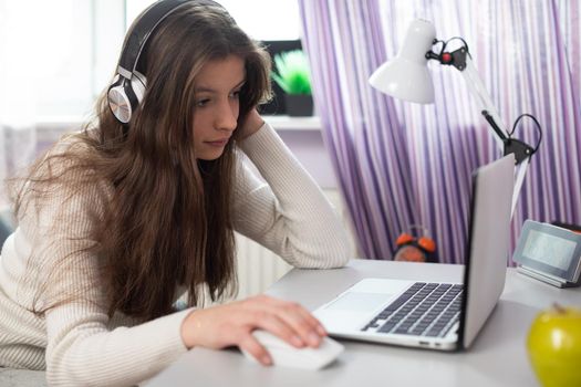 A young female student is taking schoolwork via the internet. An attractive brunette with long hair sits at a desk near a window.