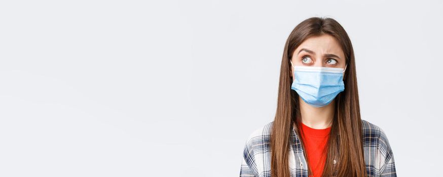 Coronavirus outbreak, leisure on quarantine, social distancing and emotions concept. Intrigued and hesitant young woman in medical mask, looking upper left corner reluctant, white background.