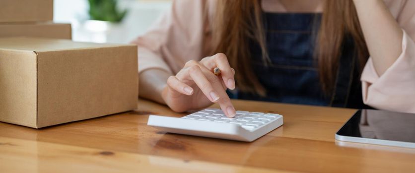 Asian woman entrepreneur using calculator with pencil in her hand, calculating financial expense at home office,online market packing box delivery,Startup successful small business owner,SME, concept.