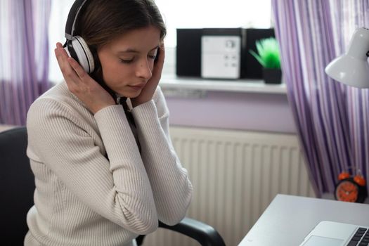 Young girl concentrates on listening to music on headphones. Attractive brunette hair styled in two braids. She sits at a desk right by the window.