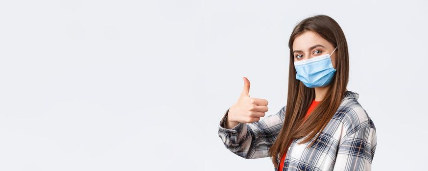 Coronavirus outbreak, leisure on quarantine, social distancing and emotions concept. Cheerful young woman in medical mask stand in profile, turn to camera and thumb-up in approval.