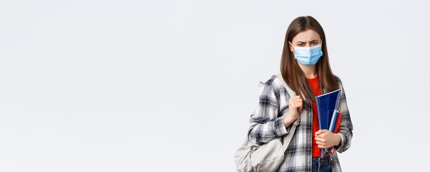 Coronavirus pandemic, covid-19 education, and back to school concept. Upset disappointed female student in medical mask hate studying in university during infection oubreak, frowning condemn dean.