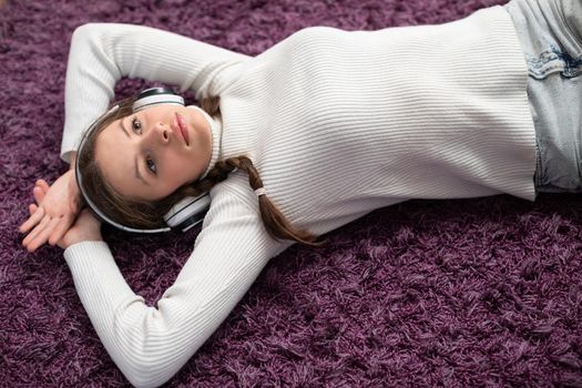 A young brunette girl is lying on the carpet listening to music and thinking about the future. View from above of pretty girl combed in two braids.