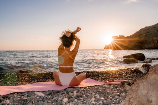 Young woman in swimsuit with long hair practicing stretching outdoors on yoga mat by the sea on a sunny day. Women's yoga fitness pilates routine. Healthy lifestyle, harmony and meditation concept.